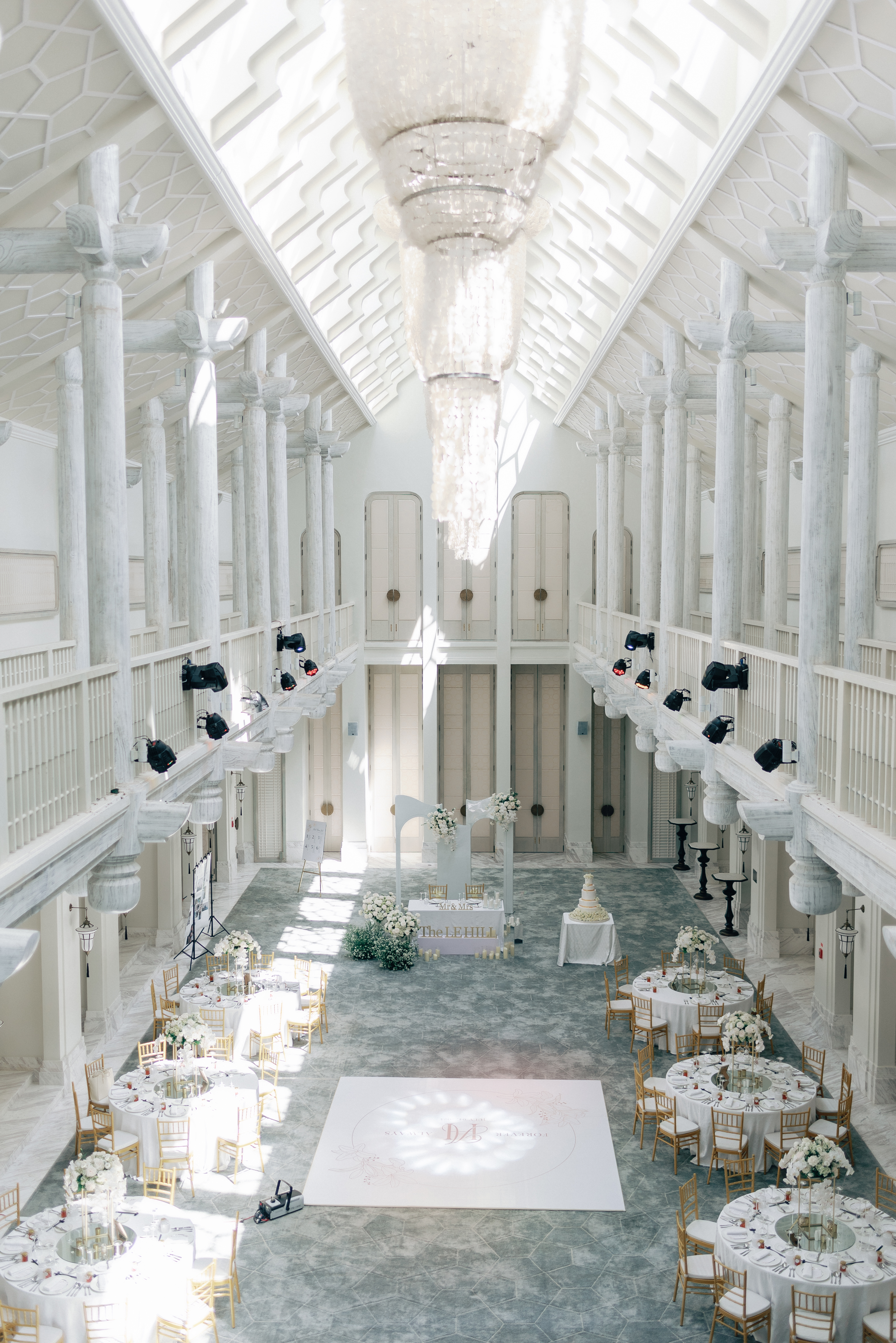Sunlit wedding reception hall with chandeliers, white floral arrangements, and gold chairs